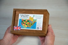 Load image into Gallery viewer, Two hands hold either side of a rectangular wooden box stained in a warm brown. atop the box is a label showing the details of the United States of America jigsaw puzzle