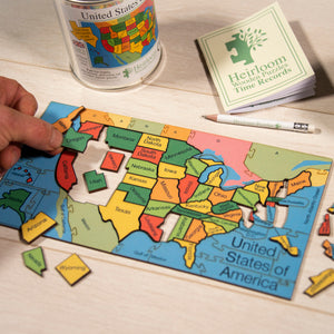 A mostly complete jigsaw of the states of america, and hand is holding and placing the piece for Oregon. Above the puzzle are the tin, pencil and Time records book.