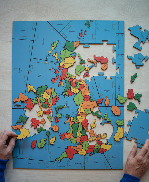 A partially made wooden jigsaw puzzle of the counties of the British Isles with two hands holding two pieces.