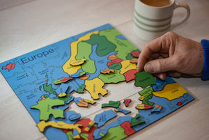A wooden puzzle of the Countries of Europe. A hand on the right of the puzzle a hand holds the Romania Piece. There is a cup of tea in the background.