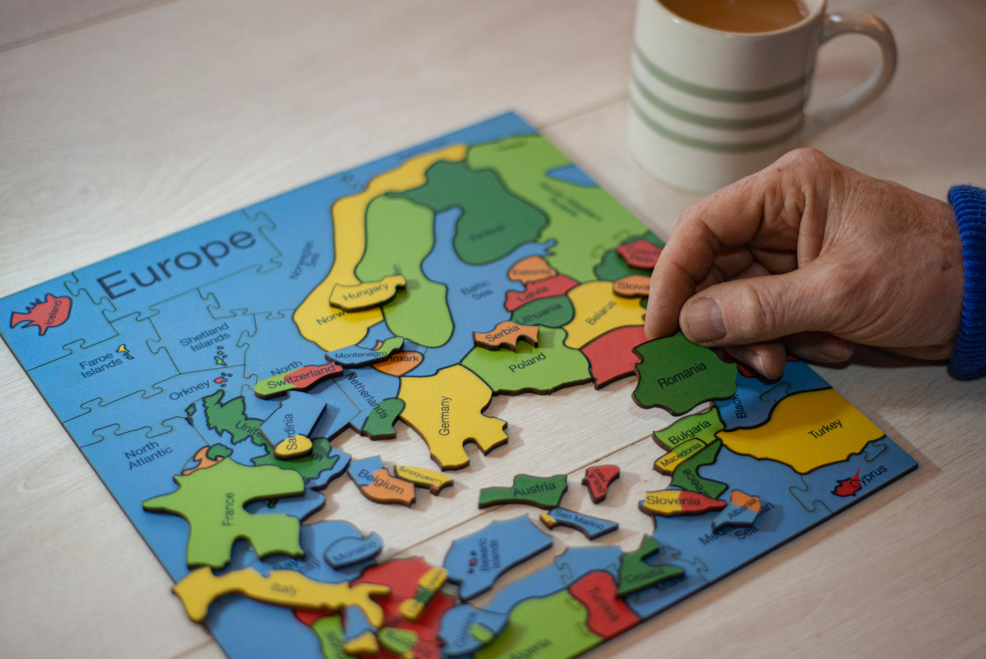 A wooden puzzle of the Countries of Europe. A hand on the right of the puzzle a hand holds the Romania Piece. There is a cup of tea in the background.