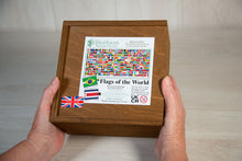 Load image into Gallery viewer, Two hands hold a warm brown stained wooden box with a sliding lid. atop the box is a label showing the details of the Flags of the world Jigsaw Puzzle