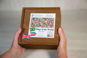 Two hands hold a warm brown stained wooden box with a sliding lid. atop the box is a label showing the details of the Flags of the world Jigsaw Puzzle